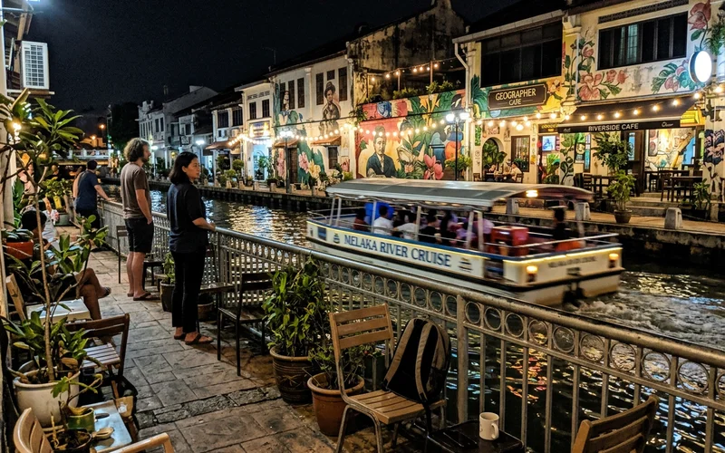 A cruise boat gliding past riverside murals and cafes after dark
