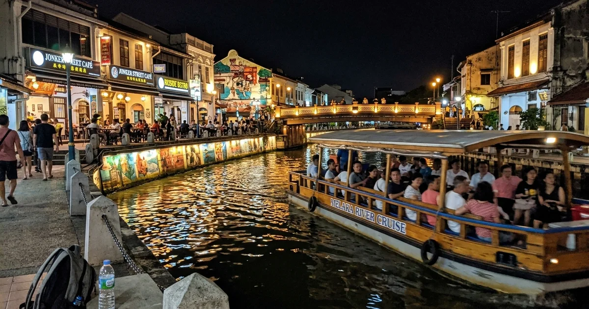 The illuminated Malacca River at night with a tour boat passing lit shophouses and a bridge