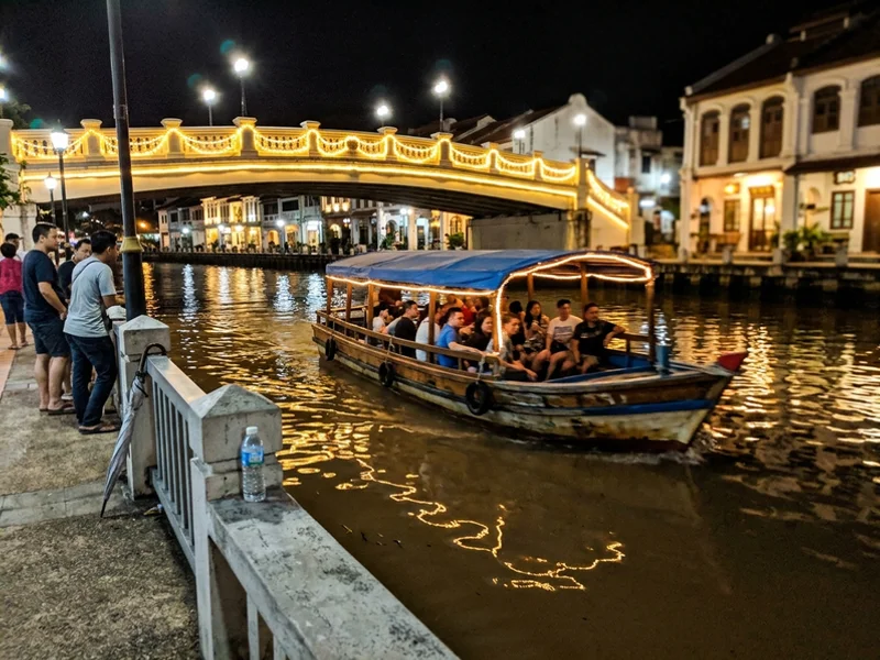 A tour boat passing under a lit bridge on the Malacca River at night